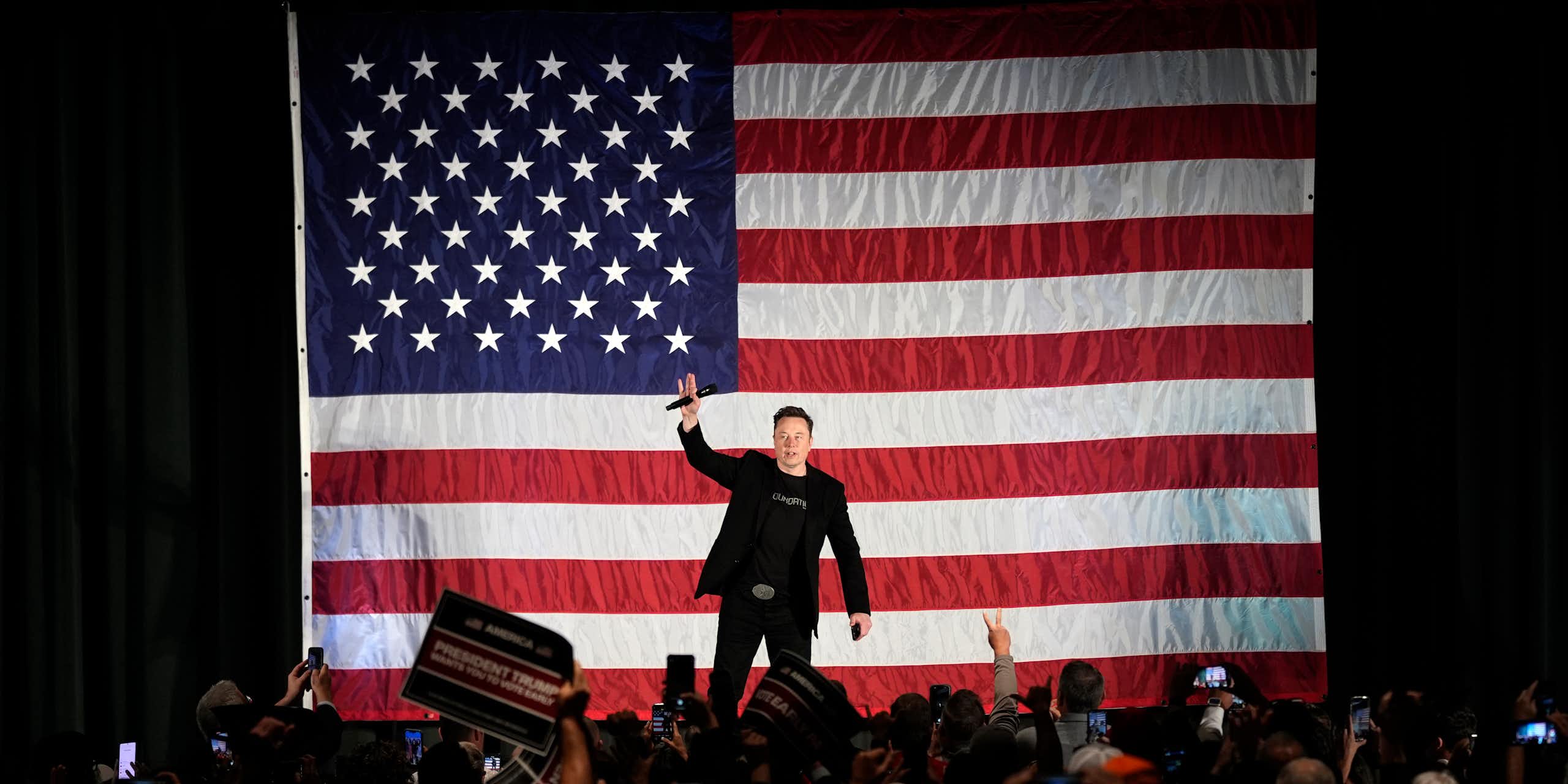 Crowd of people taking photos of a man on stage holding a microphone in front of a large American flag.
