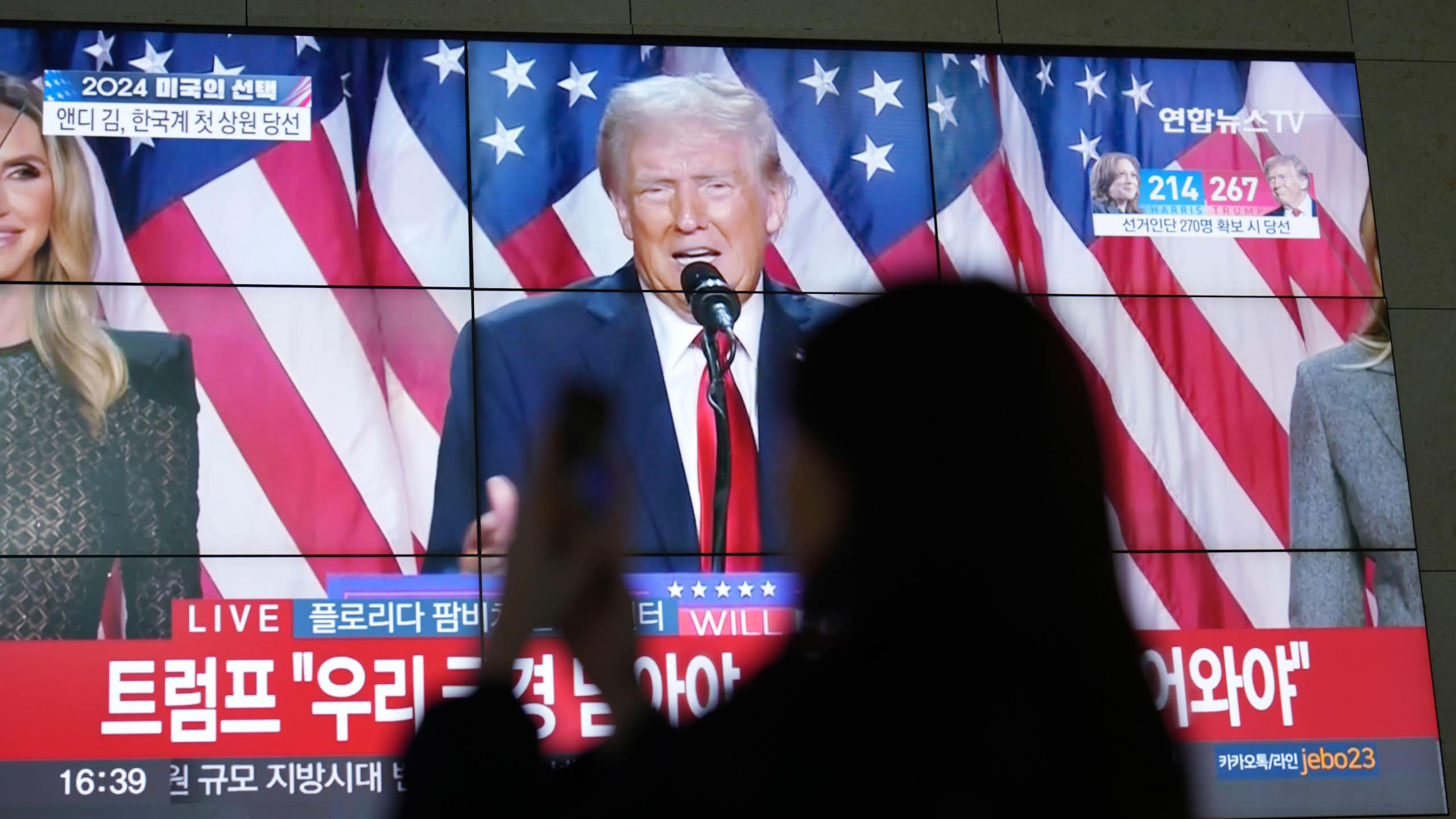 A silhouette of a woman looking at her phone with Donald Trump on a TV screen behind her.