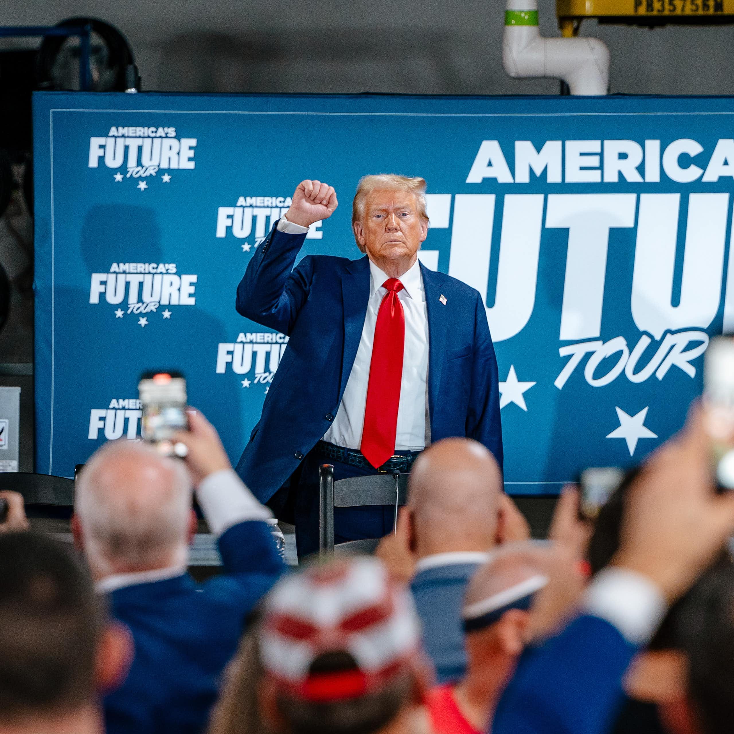US Republican presidential candidate Donald J. Trump raises his fist for a crowd in a factory, in front of a banner that reads America's Future Tour
